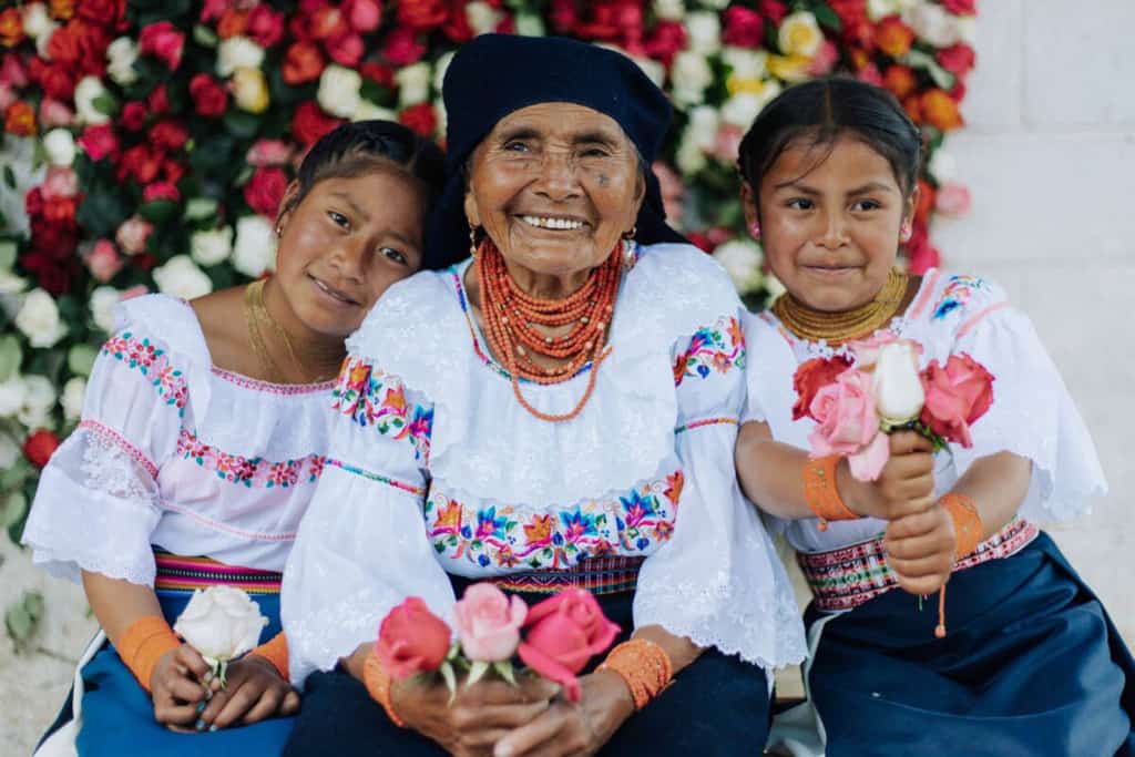 Angelita is sitting in the middle of her two granddaughters. They are all wearing traditional clothing and are holding roses. Behind them is a wall of roses.