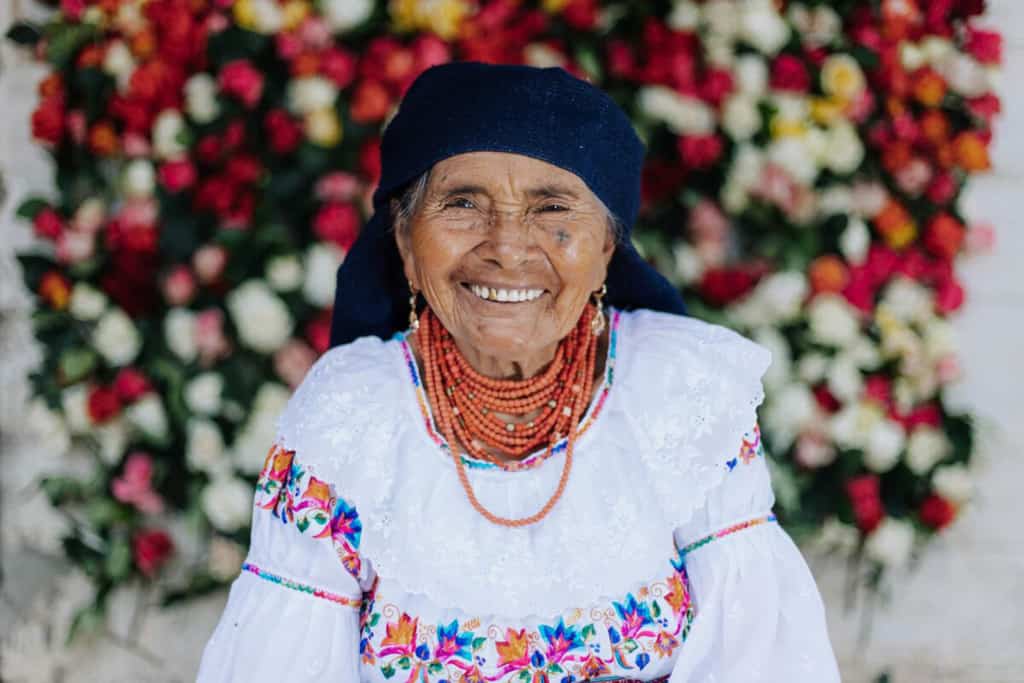 Angelita is sitting at her home with a wall of roses behind her. She is wearing traditional clothing.