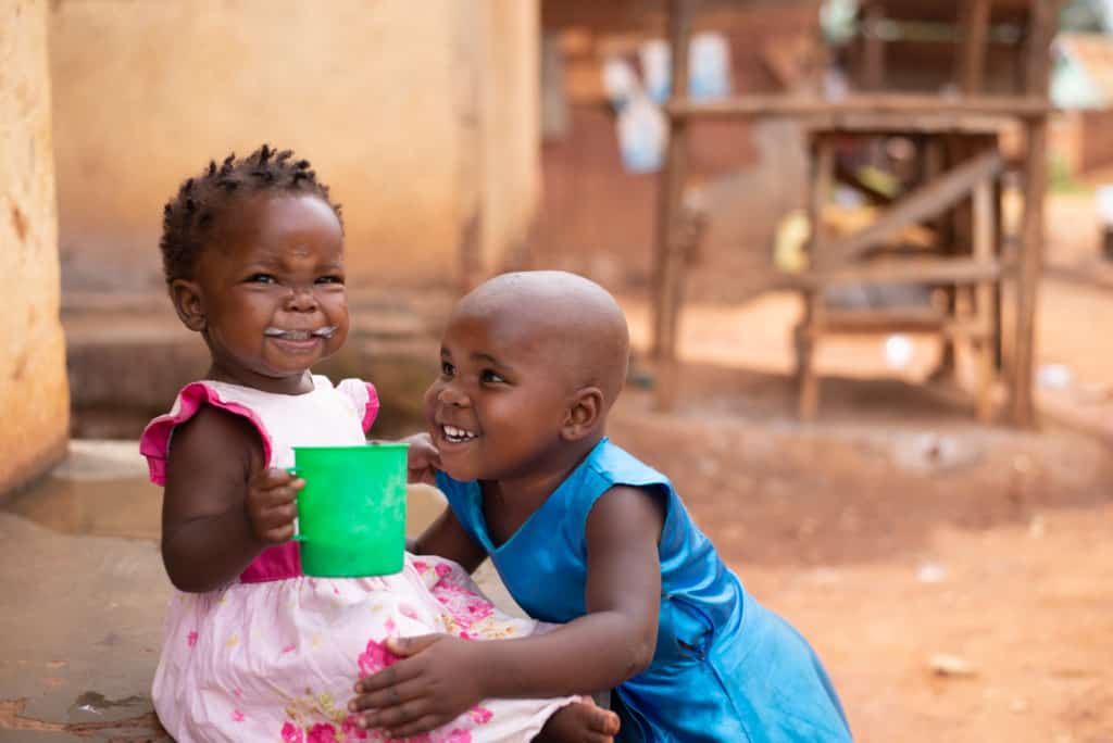 A toddler smiles with porridge on her face. She is wearing a pink dress and holding a green cup of porridge. Her sister is smiling next to her, wearing a blue dress.