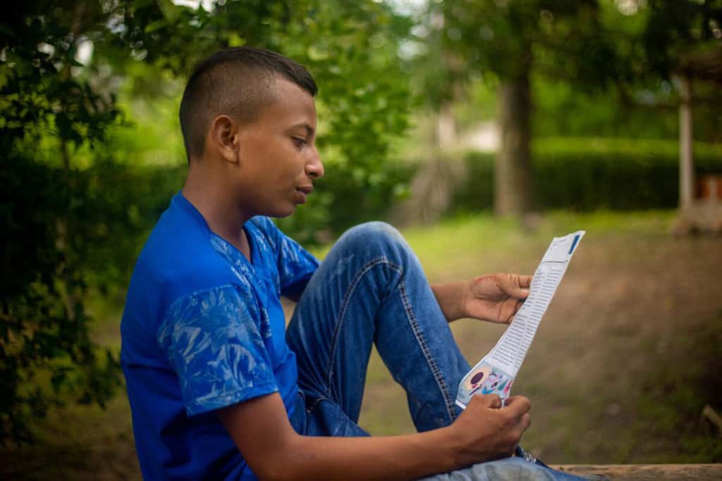 Carlos is wearing jeans and a blue shirt. He is sitting down outside reading one of his sponsor's letters. There are trees in the background.