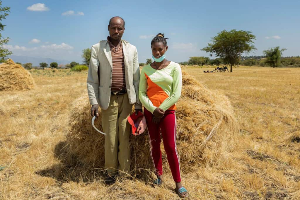 Dagem is wearing red pants, an orange and green shirt, and a face mask. She is standing next to her father in a field where they are harvesting crops.