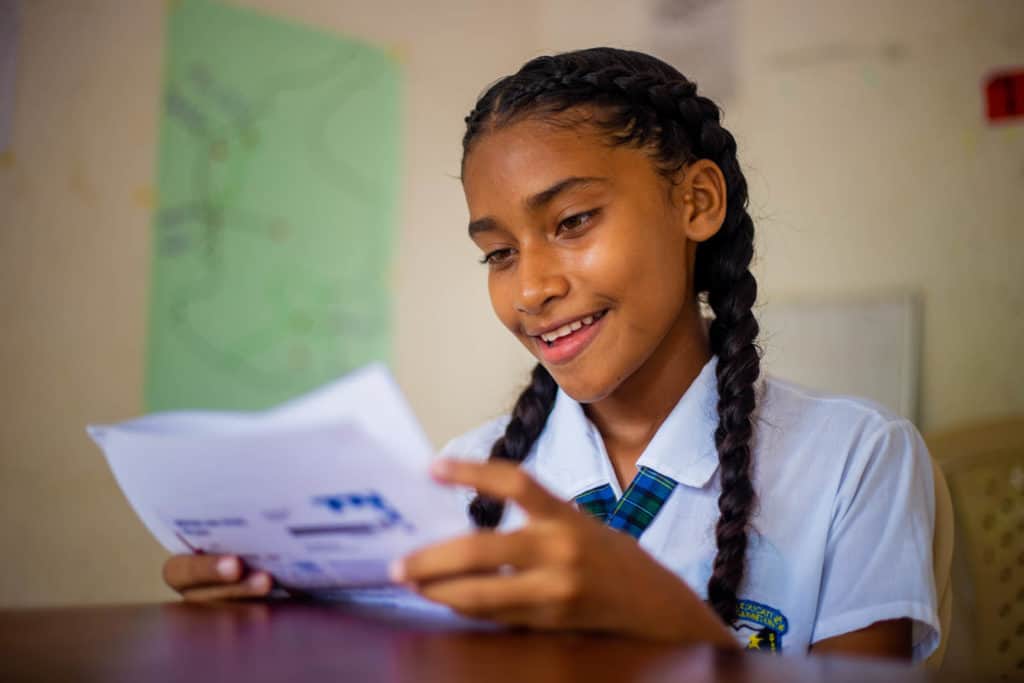 Sara, in a white uniform shirt, is sitting at a table at the project. She smiles while reading a letter she received from her sponsor Pamela. 