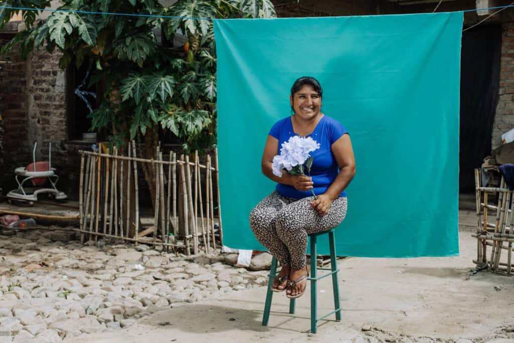 Yhovana is wearing a blue shirt with black and white pants. She is sitting on a stool in her backyard and is holding flowers. Behind her is a green backdrop.