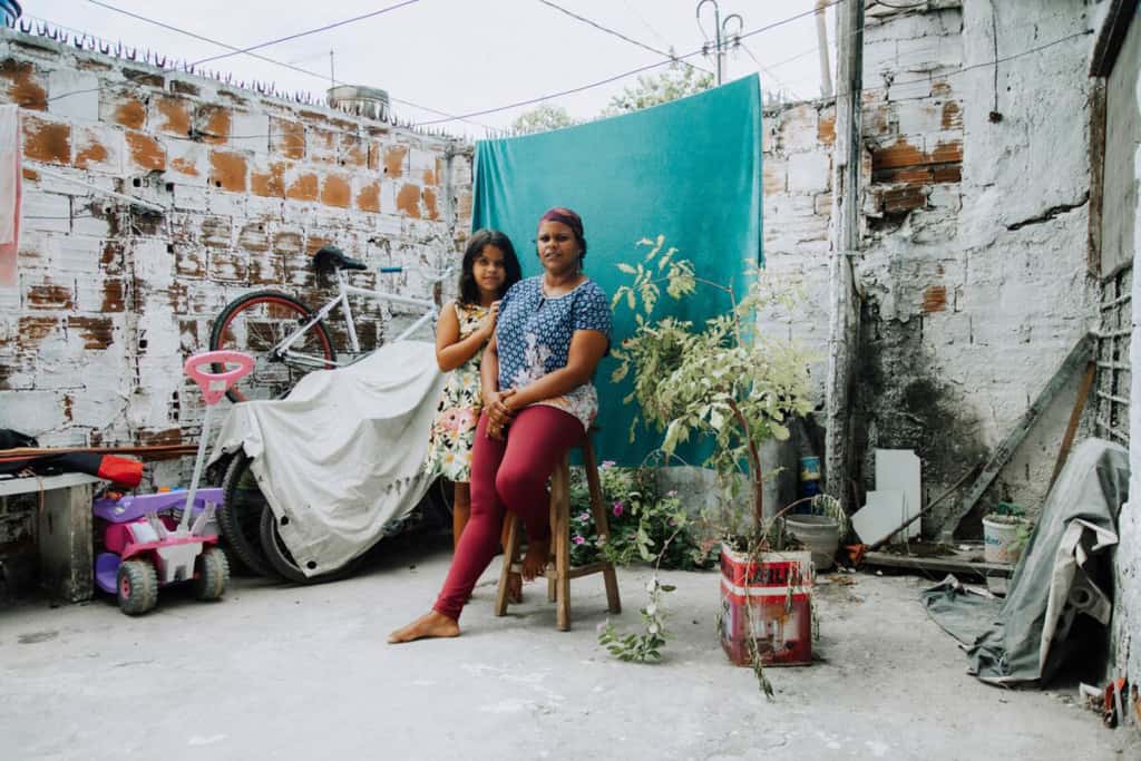 Danielly is wearing a blue shirt and maroon pants. She is sitting on a stool outside her home in front of a green background. Her daughter, Rayane, is standing next to her.