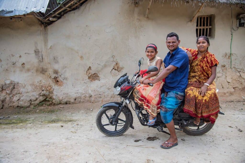 A young girl sits on the front of a motorcycle. Behind her is her father and her mother. They are wearing colorful clothing.