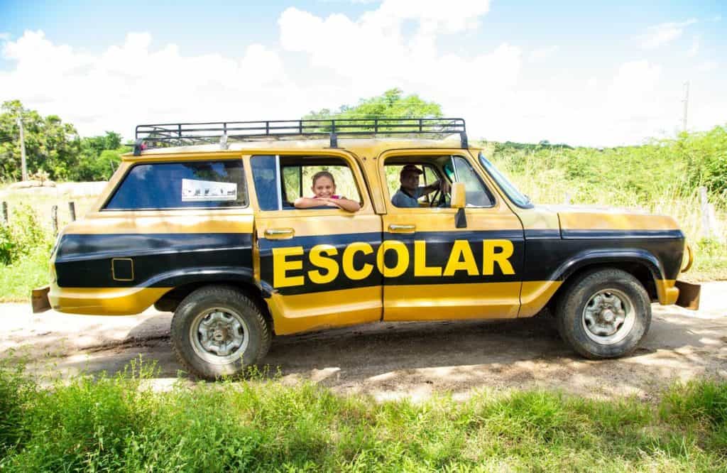 A black and yellow jeep truck vehicle reading "Escolar" is parked on a dirt road. A smiling girl looks out the window.