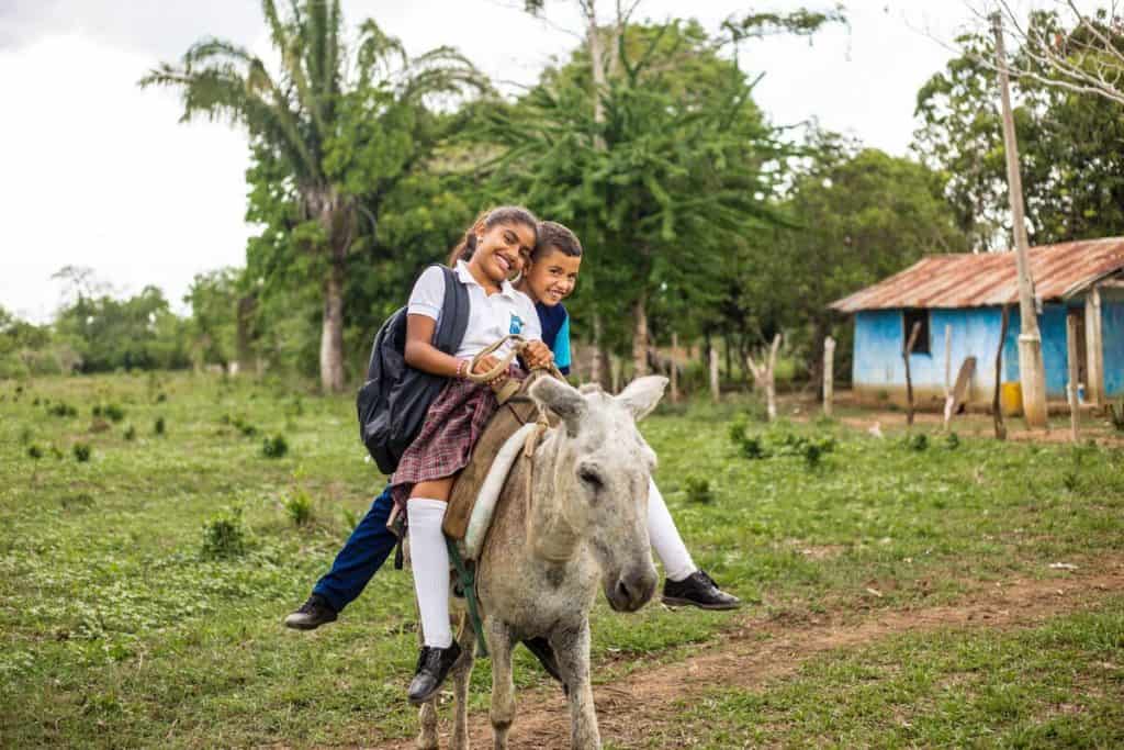 A brother and sister ride on a donkey. The girl is wearing a school uniform and backpack.