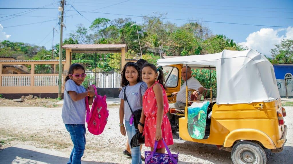 Three girls, friends, are getting ready to board a yellow tuk-tuk vehicle, which a man is driving.