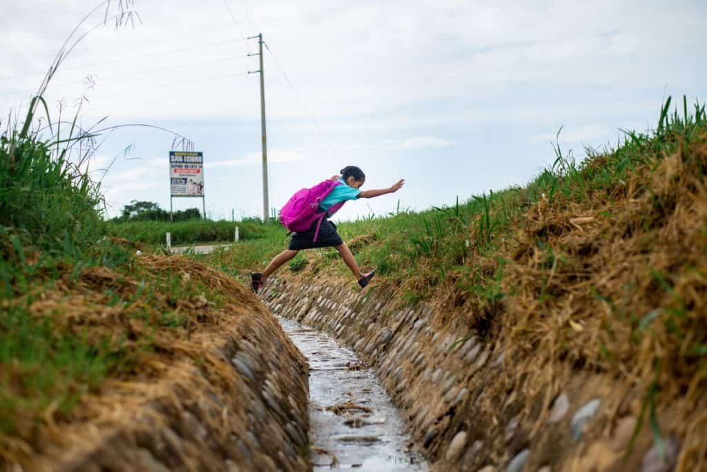 A girl wearing a blue shirt and pink backpack jumps across a ditch. There is water running down the ditch.