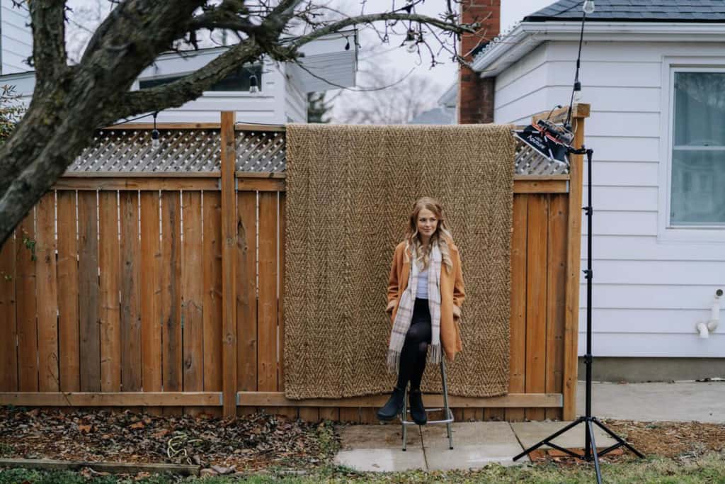 Becky is sitting on a stool in her back yard. Behind her is a wooden privacy fence with a brown rug hanging over it.