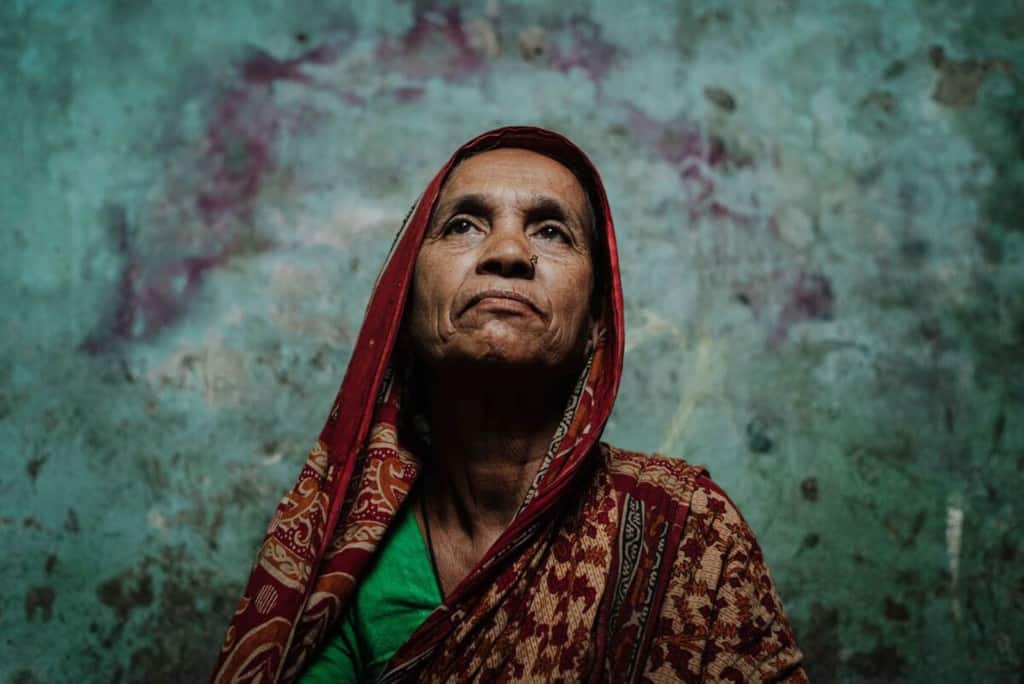 Woman wearing a red and tan dress. She is sitting inside her home in front of a green wall and she is looking upward.