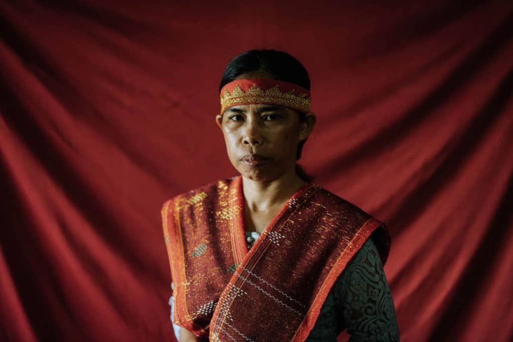 Marhati is sitting in her home in front of a red backdrop. She is wearing traditionally woven clothing.