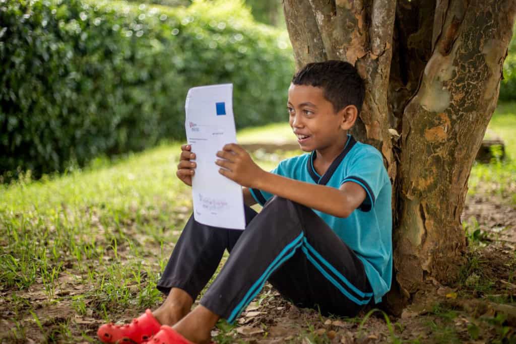 Carlos is wearing a blue shirt and black pants. He is outside, sitting against a tree, and reading a letter from his sponsor.