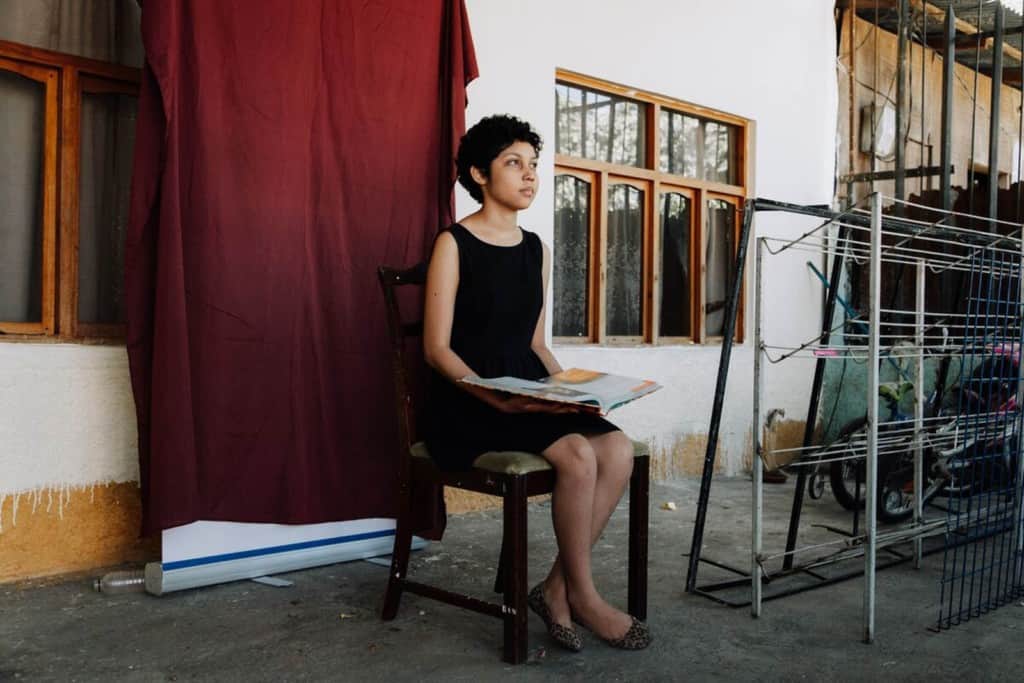 Diana is wearing a black dress. She is sitting in a chair in front of a red backdrop in her home and is holding a book.