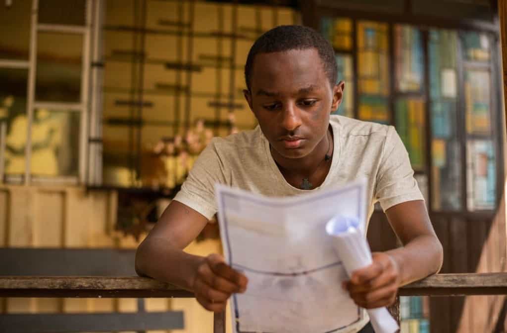 Adonay is wearing a gray shirt and is standing outside reading a letter from his sponsor.