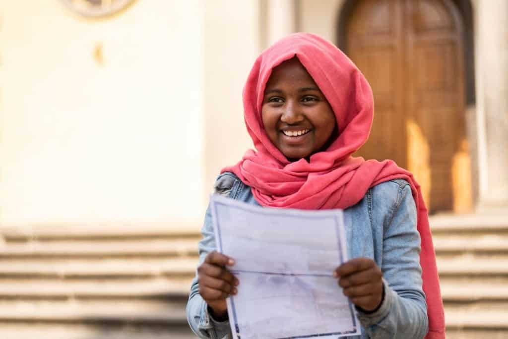Liya is wearing a jean jacket and a pink head covering. She is standing outside and is holding a letter from her sponsor.
