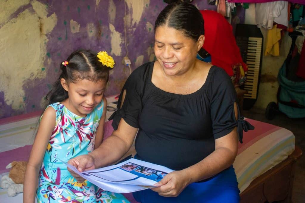 Abigail is wearing a blue dress with a bright floral pattern. She is sitting on her bed with her mother, Claudia. They are reading one of Abigail's sponsor letters.