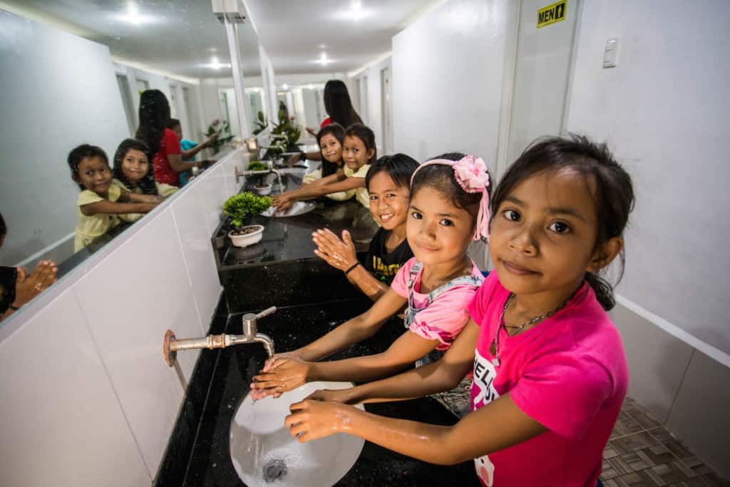 Several girls are seen here in the bathroom at the Student Center. They are smiling and looking at the camera as they wash their hands.