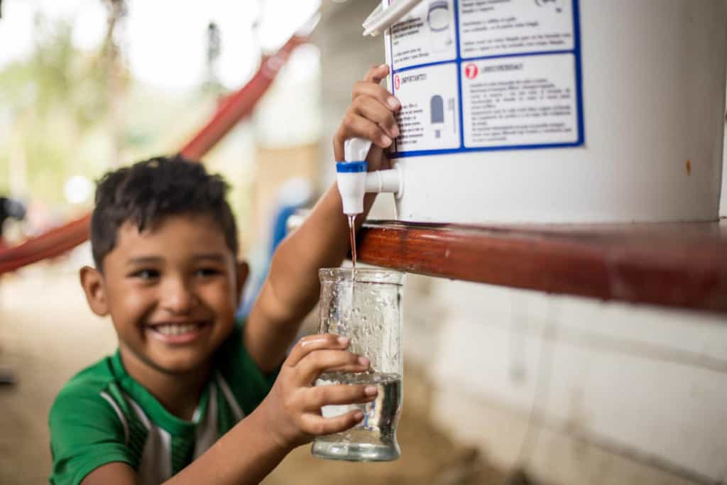 Boy taking water to drink from the water filter he received from the project. He is wearing a green tee shirt.