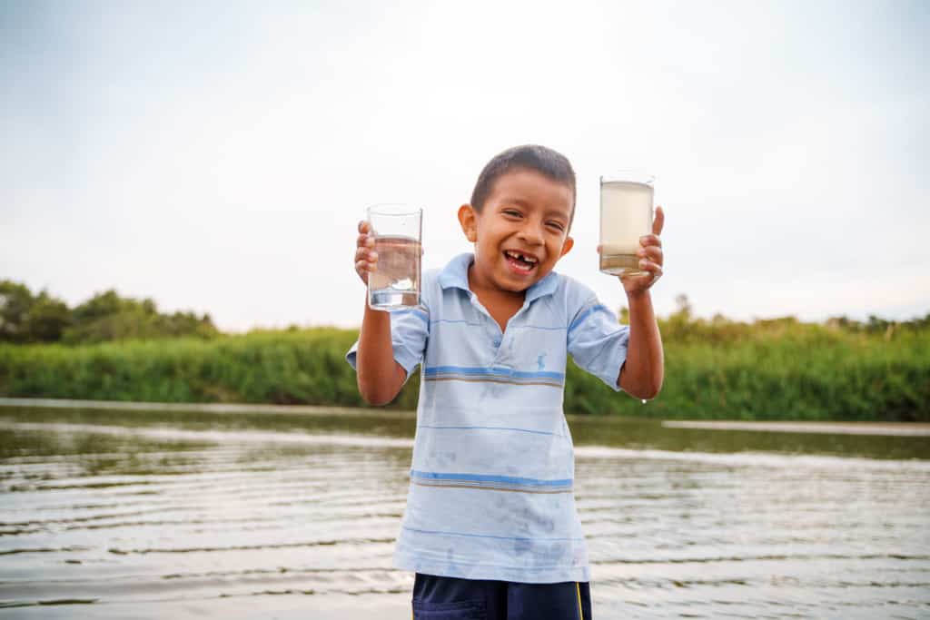 A boy wearing a light blue shirt is standing in front of a body of water. He is holding a glass of water in each hand. One glass contains clean water and one contains dirty water. There are trees behind the water.