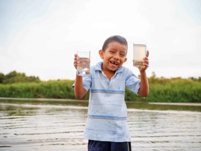 A boy wearing a light blue shirt is standing in front of a body of water. He is holding a glass of water in each hand. One glass contains clean water and one contains dirty water. There are trees behind the water.