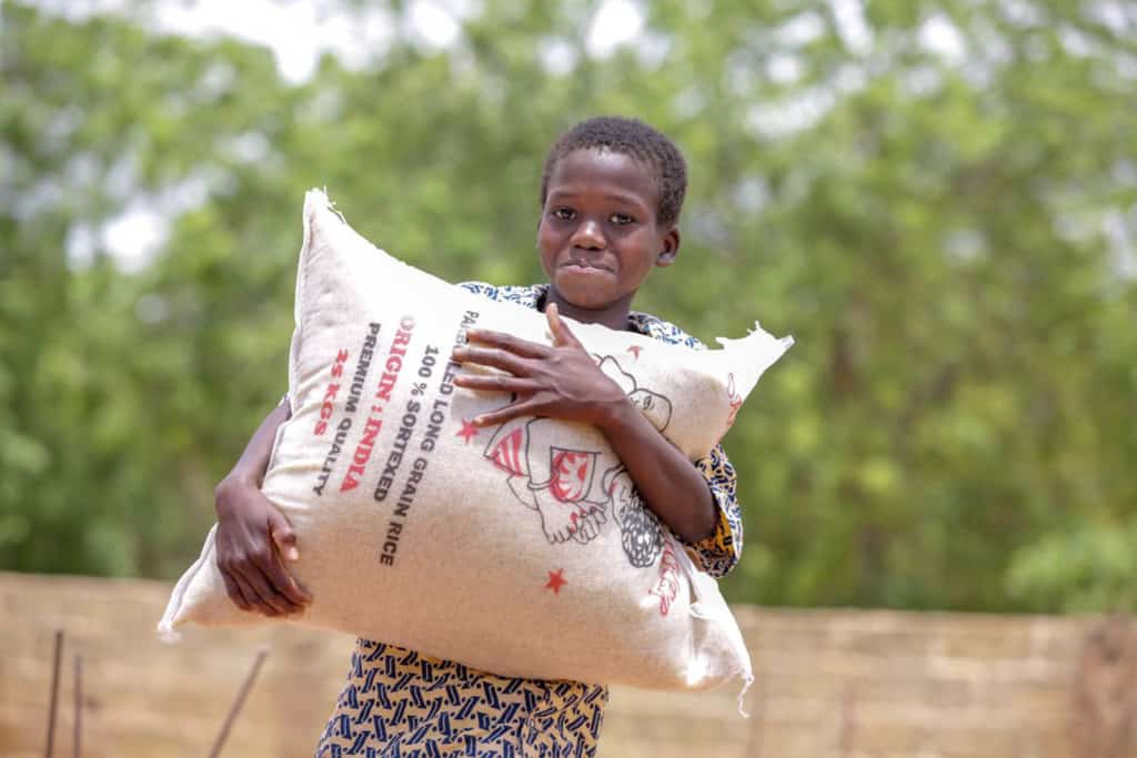 A child is seen here smiling at the camera and carrying a large bag of food. There is a brick wall and trees in the background.