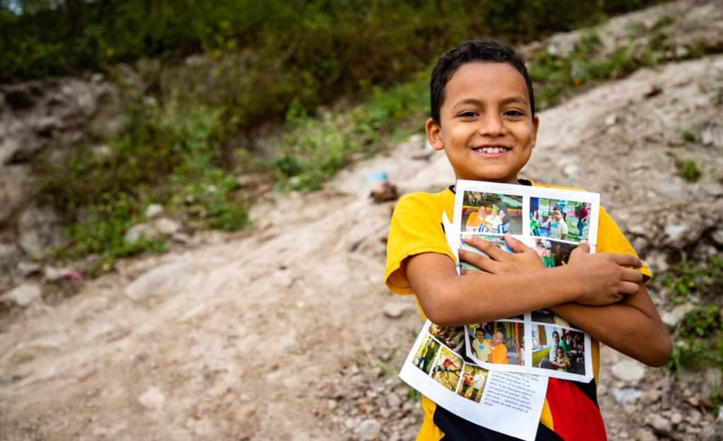 Boy wearing a yellow shirt and jeans. He is standing outside his home and is holding his sponsor letters close to his chest.