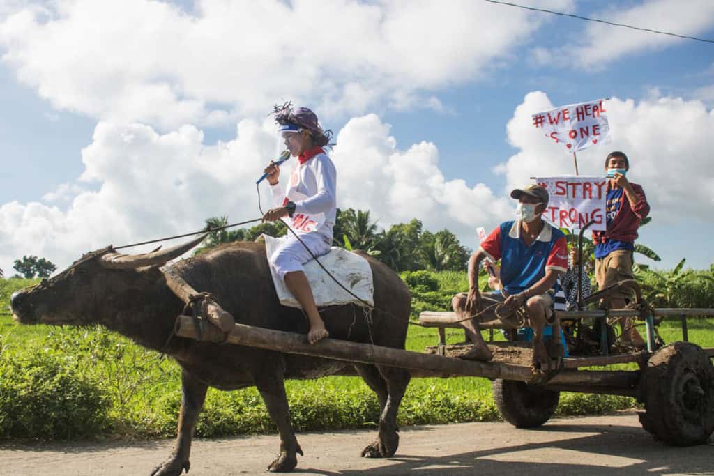 A boy and his carabao (water buffalo) karaoke challenge. They are walking down a road and people are holding signs as they sit in a cart pulled by the water buffalo.