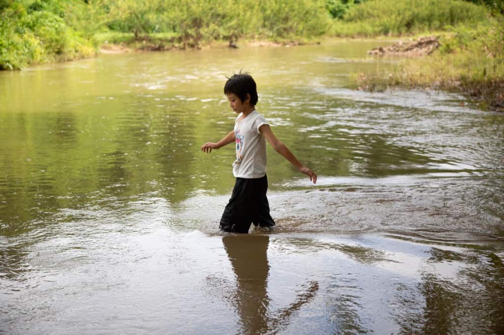 Girl wearing a white shirt and black shorts. She is walking into a stream near her village.