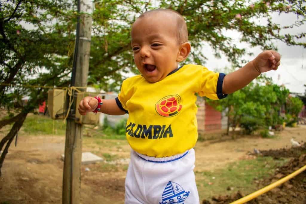 Baby Deinner is wearing a yellow Colombia football shirt and white pants with a sailboat on them. He is smiling and outdoors