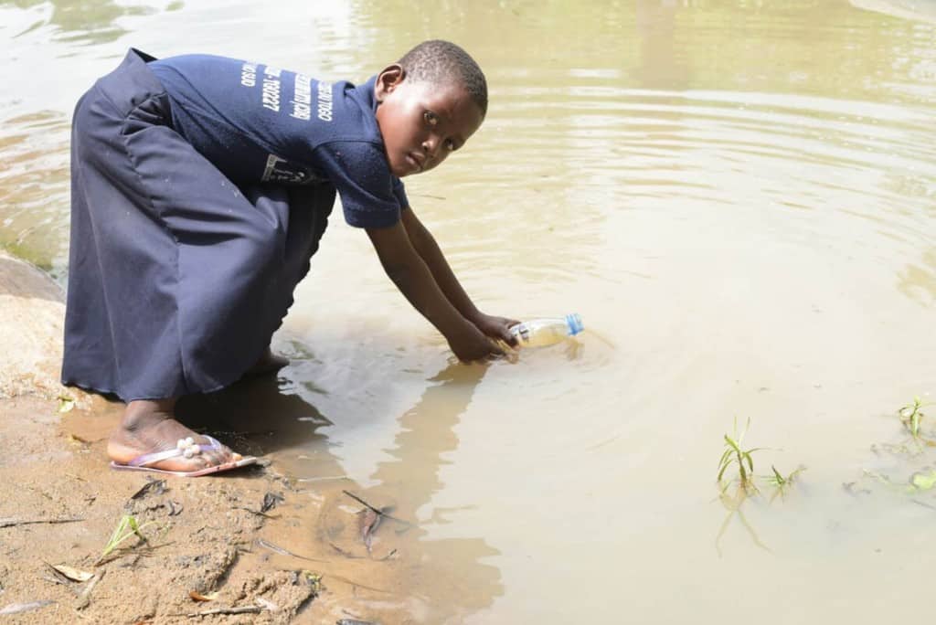 Girl wearing her Compassion uniform, a blue shirt and skirt. She is standing at the edge of the river where they gather water.