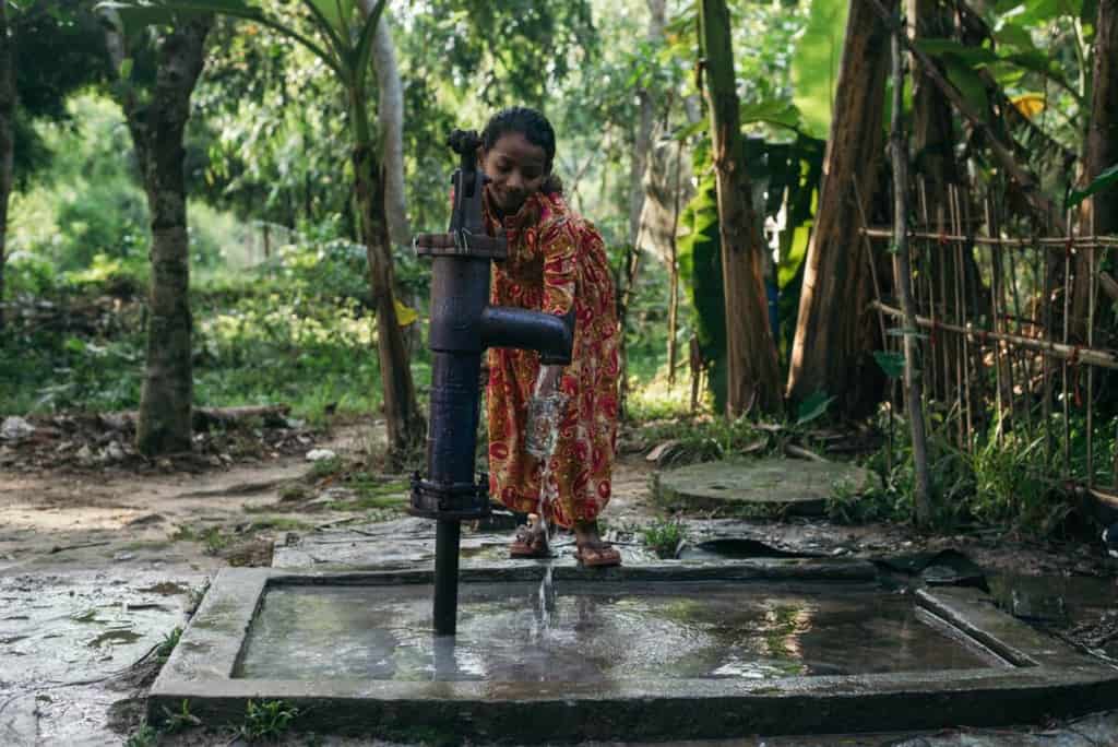 Girl wearing a gold dress with a red paisley pattern. She is pumping water from the tube well at her home. There are trees behind her.
