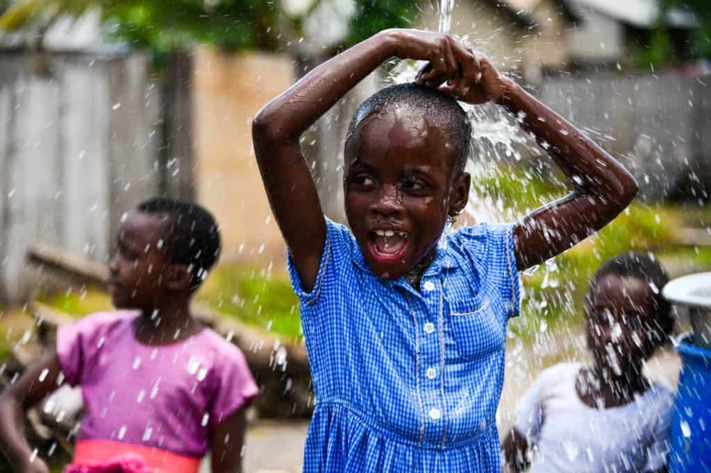 Girl wearing a blue and white checked dress. She is playing in water coming from the pipe to the borehole at the Compassion center. The water is running down over her head. Other children are in the background.