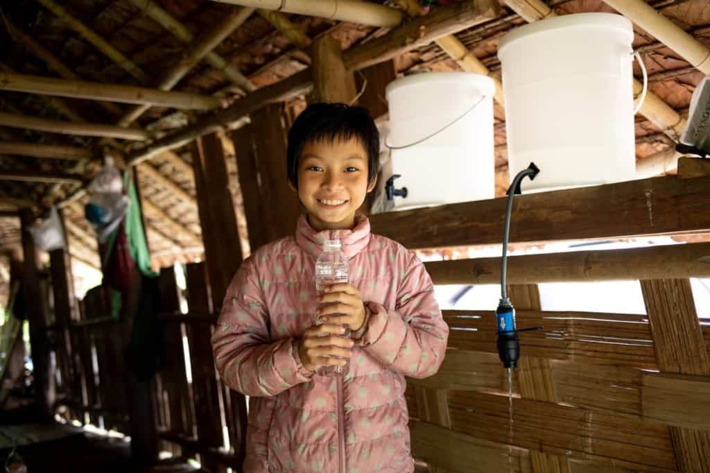 Girl wearing a pink jacket. She is inside her home and is holding a water bottle full of clean water.