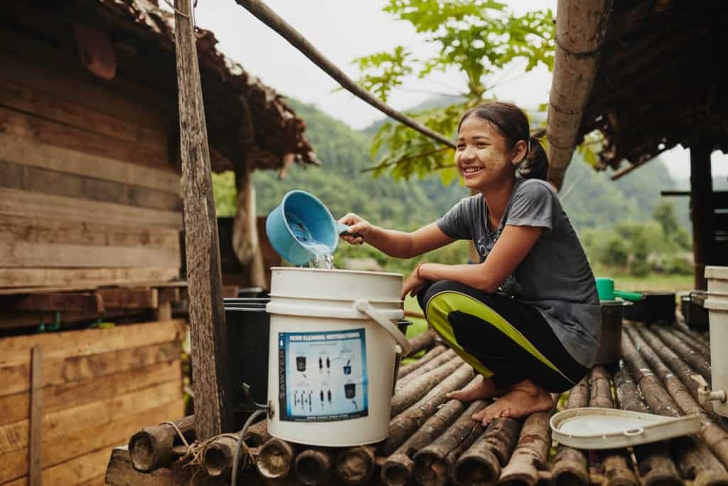 A smiling girl in gray shirt black and green pants squats, sits on a bamboo floor next to a white bucket, pouring water into it from a light blue pan.
