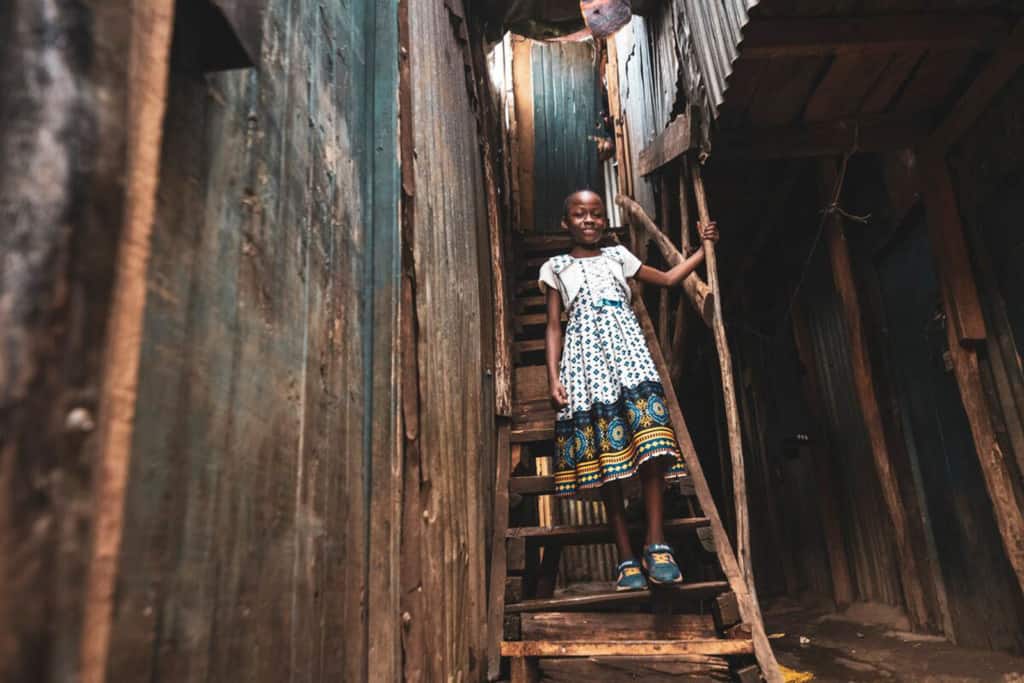 Girl wearing a white dress with a blue pattern. She is standing on a wooden stairway between two houses in the slums, where she lives with her family.