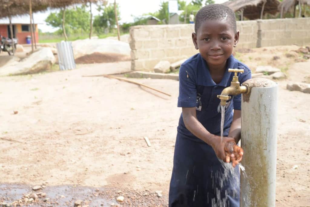 Girl standing at a faucet and is washing her hands. She is wearing a blue shirt and skirt.