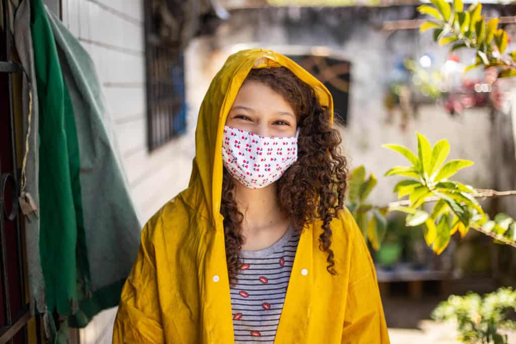 Girl standing in front of her home. She is wearing a black and gray striped shirt, yellow raincoat, and a face mask.