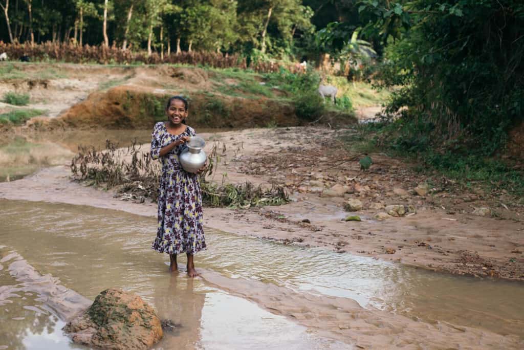 Girl wearing a white dress with a floral pattern. She is holding a silver jug and is walking down the shallow riverbed.