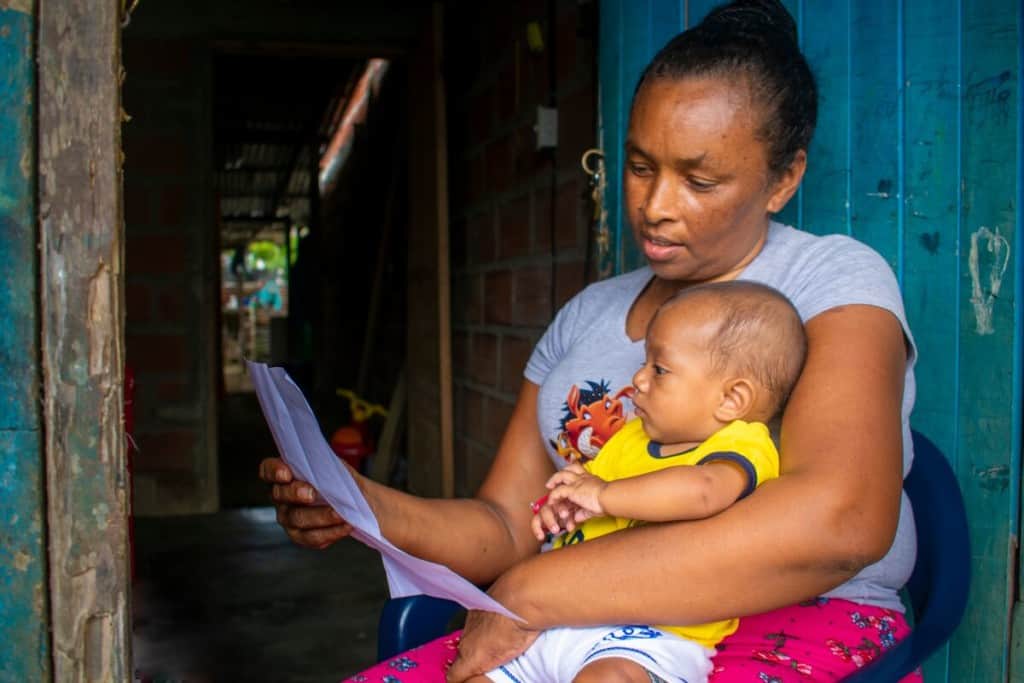 A grandmother is holding her newborn grandson and reading from a sheet of paper.