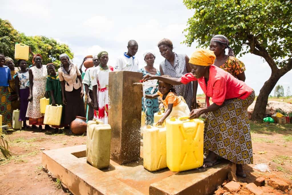 Woman wearing a yellow head scarf and a red shirt with a long pattern skirt smiles as she fetches clean water from an outdoor outside community faucet into her yellow plastic container Other women standing single file in line behind her to also fetch the clean running water. Girl wearing an orange and white dress helps her mother collect the life saving water.