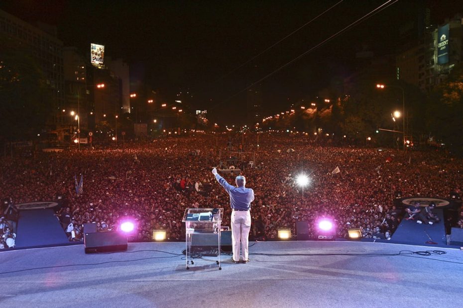 Luis Palau preaching to a huge audience in Buenos Aires. He is standing on a stage holding a hand in the air and wearing a blue shirt and white pants.