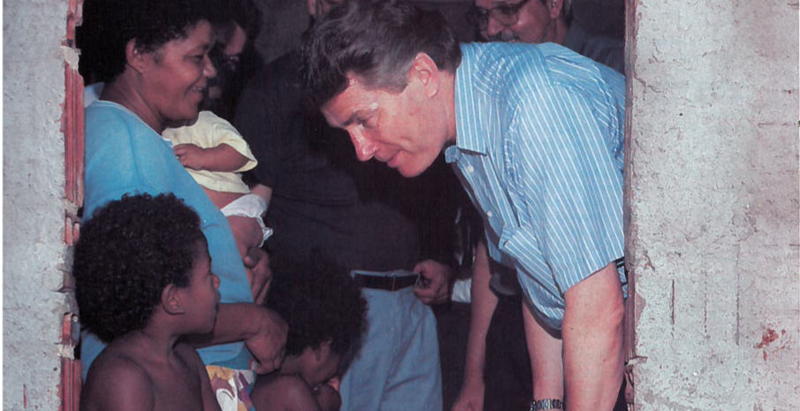 Luis Palau bends down to speak to a child. He is wearing a blue and white striped shirt.