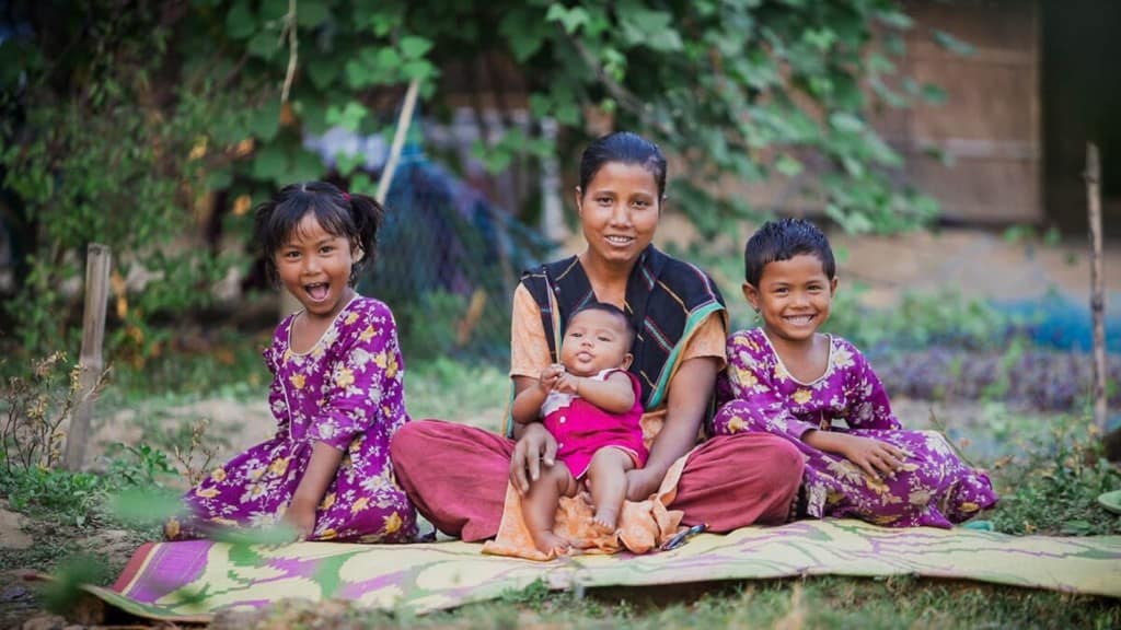 Mother and children  all sitting together on a blanket outside their home. Two of the chilren are wearing dark pink dresses with flowers on them.