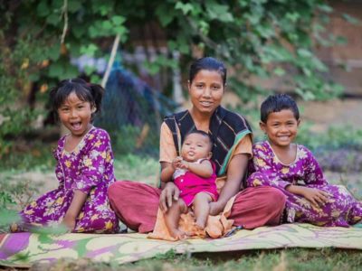 A woman and her three children sit on a mat outdoors. They are smiling. The two older children are wearing matching purple dresses