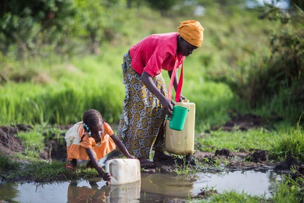 Woman wearing a yellow scarf on her head, a red shirt and long pattern skirt at a large puddle where rain water collected on the ground.