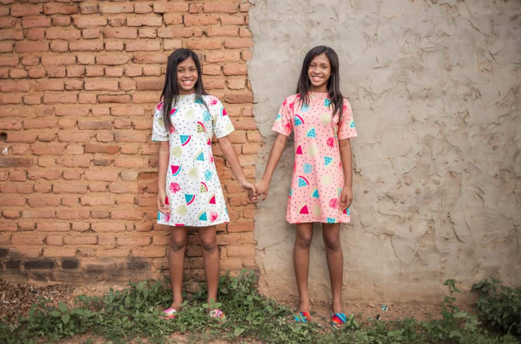 Sarah is wearing a pink patterned dress and Sophia is wearing a white patterned dress. They are standing in front of a brick and cement building and they are holding hands.