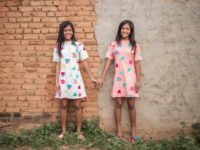 Sarah is wearing a pink patterned dress and Sophia is wearing a white patterned dress. They are standing in front of a brick and cement building and they are holding hands.