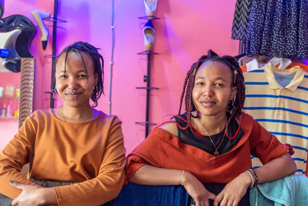 Miriam is wearing a light orange shirt and Maureen is wearing a dark orange shirt. They are inside their salon and behind them, clothes are hanging on the wall.