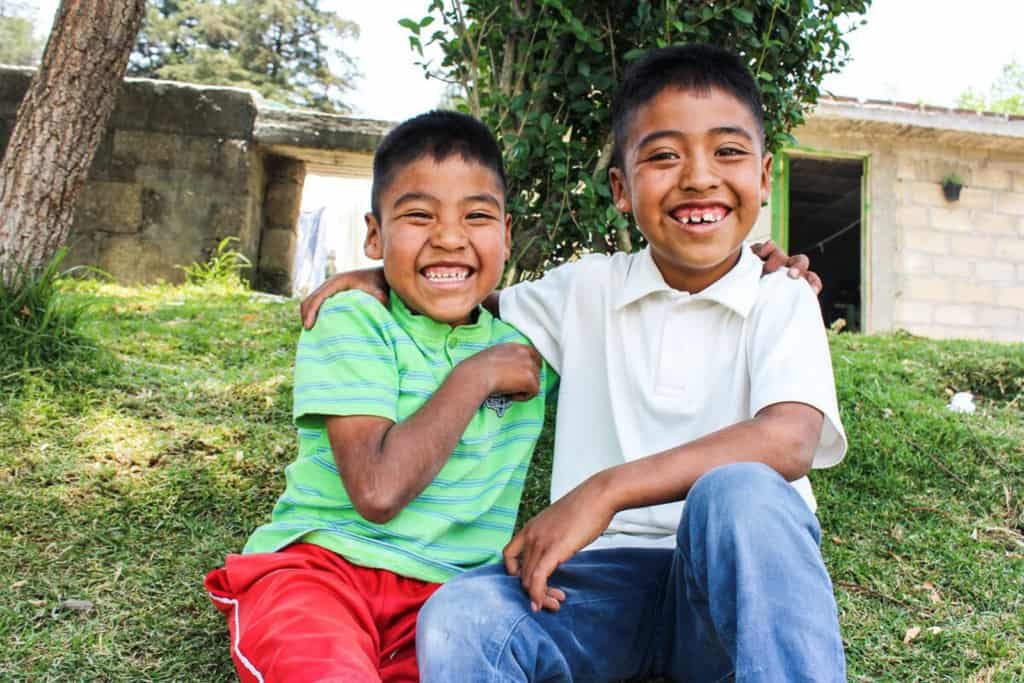 Jose, wearing a green shirt and red shorts, is sitting outside his home with his brother, Victor, wearing a white shirt and jeans. They have their arms around each other.